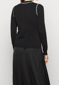 Femme portant un pull à manches longues noir avec un liseré blanc "MK" sur les épaules et le col, associé à une jupe noire plissée.