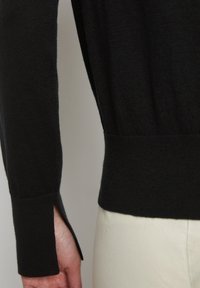 Czarny dziergany sweter z długimi rękawami i bocznymi rozcięciami przy mankietach, z subtelną teksturą i dopasowanym pasem, zestawiony z jasnymi spodniami.