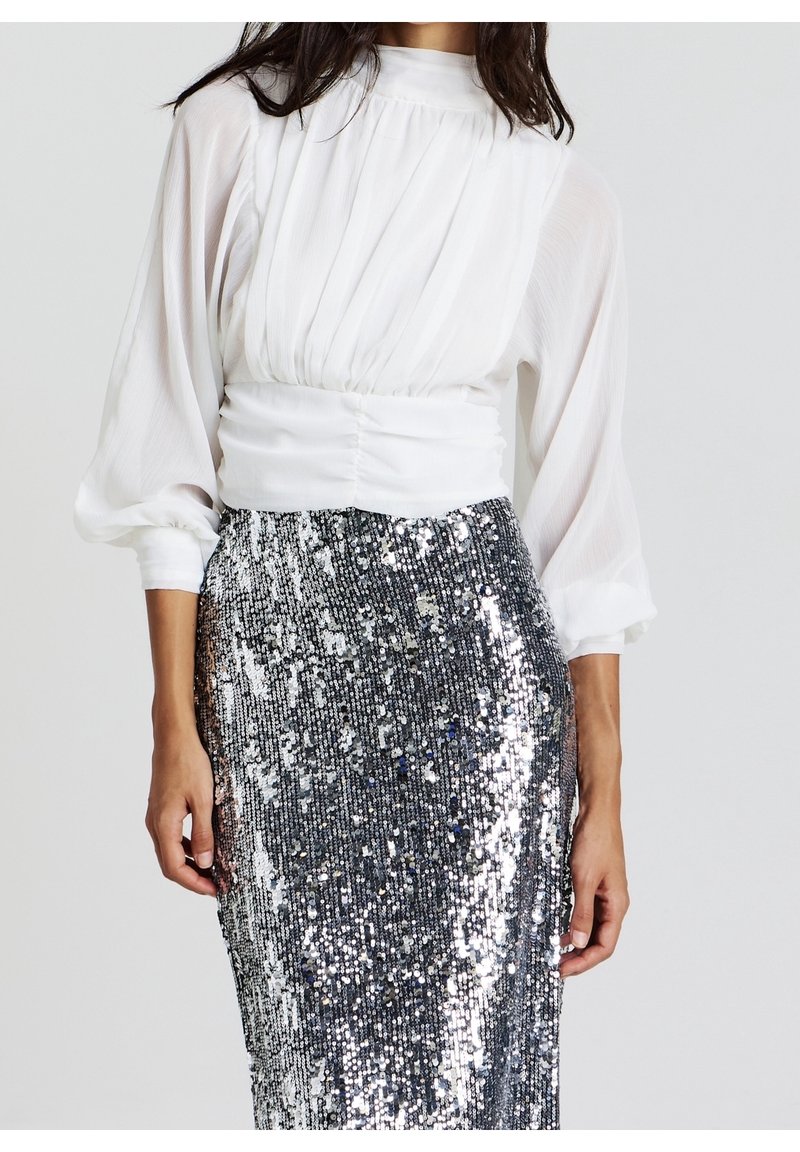 Femme portant un chemisier blanc à manches longues avec des détails froncés et une jupe argentée à sequins taille haute sur un fond uni.