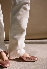 Błyszczące baletki w kolorze różowego złota z okrągłym noskiem i niskim obcasem, w połączeniu z białymi spodniami o szerokich nogawkach na betonowej powierzchni.