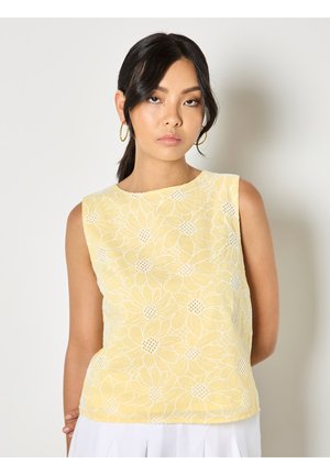 Femme aux cheveux foncés attachés en queue de cheval, portant des boucles d'oreilles créoles dorées et un haut jaune sans manches avec un motif floral blanc, debout devant un fond uni.