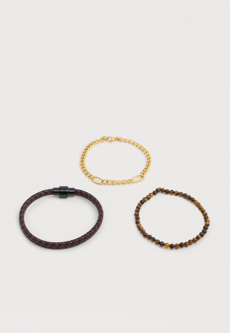 Trois bracelets : chaîne en or, perles marron, et cuir tressé rouge foncé et noir avec fermoir noir sur fond blanc.