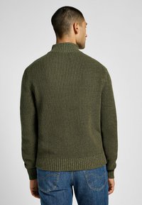 Tumma vihreä neulepusero korkealla kauluksella, ribbipinnalla ja istuvilla hihoilla. Yhdistetty sinisiin farkkuihin, näyttäen takanäkymän.