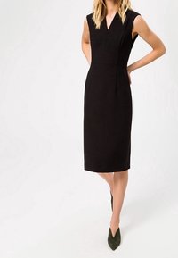 Svart ärmlös klänning med V-hals, knälång, figurnära silhuett, slät tyg och inga synliga mönster; matchad med gröna spetsiga klackar.