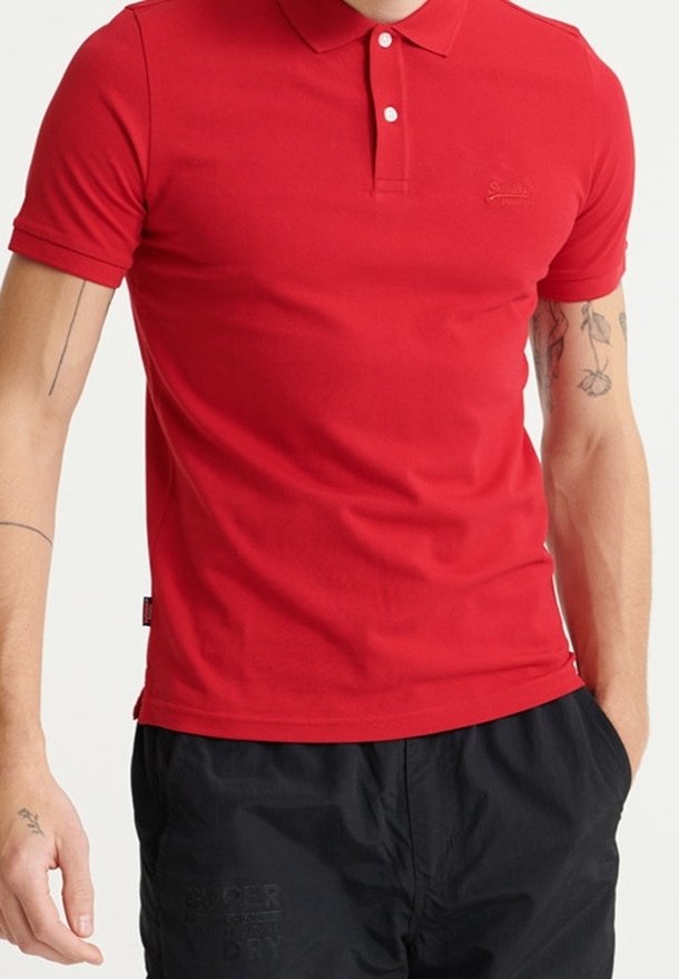 Homme portant un polo rouge ajusté à manches courtes et un pantalon noir avec des tatouages visibles sur les bras sur un fond clair.