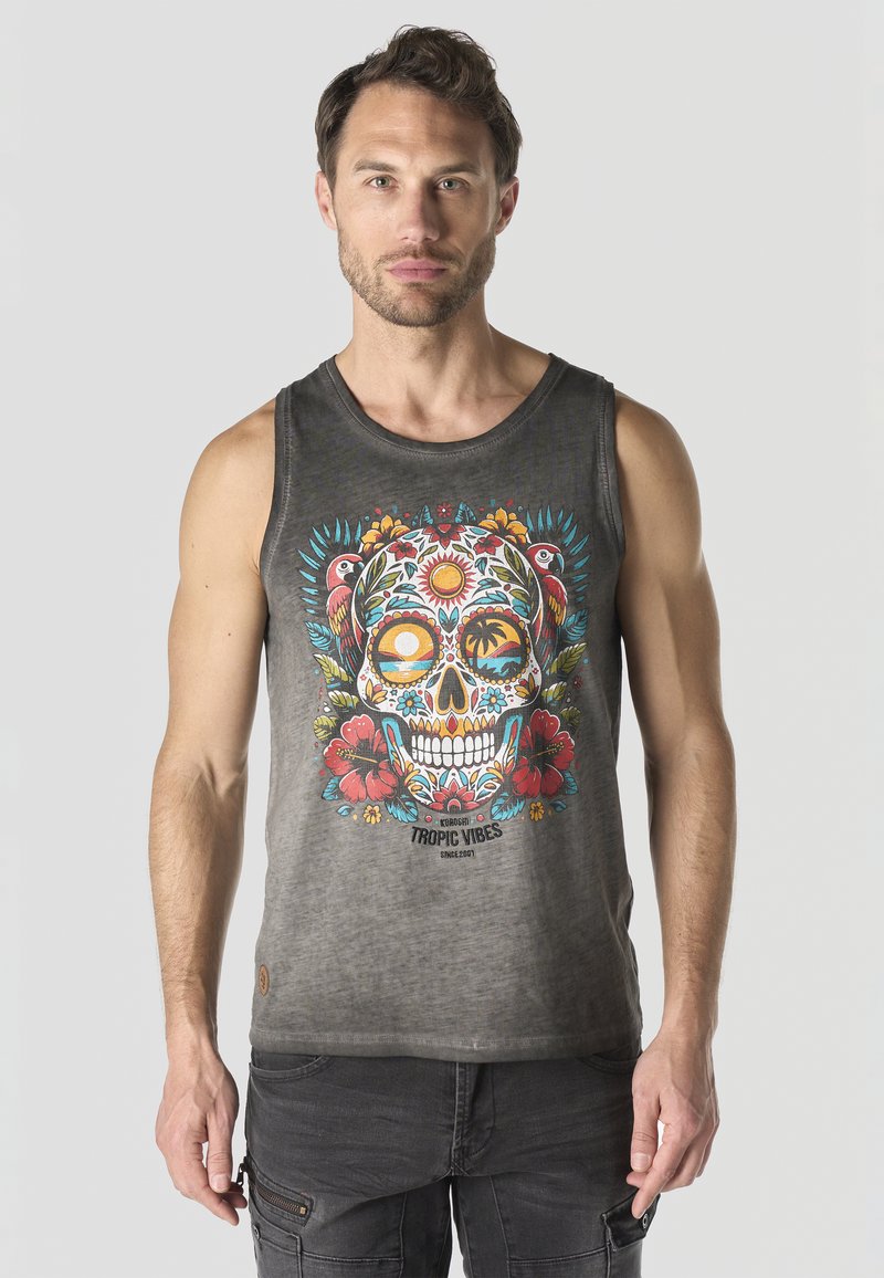 Hombre que lleva una camiseta sin mangas gris con calavera de azúcar colorida, loros, flores y texto "Tropic Vibes" de pie contra un fondo neutro.