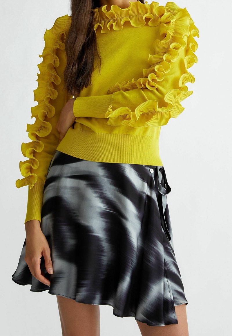 Femme portant un pull jaune avec des détails froncés aux manches et à l'encolure, associé à une jupe à motif abstrait noir et blanc.