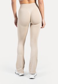 Beżowe legginsy z wysokim stanem o gładkiej fakturze, rozszerzanych nogawkach i dopasowanym kroju. Noszone z białymi butami sportowymi.