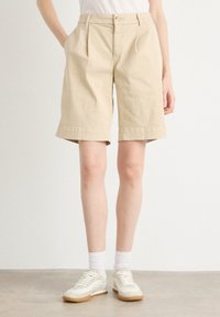 Shorts en coton beige avec des plis, des ourlets retroussés, des poches latérales et une fermeture à bouton à l'avant, portés avec des baskets blanches et des chaussettes de cheville.