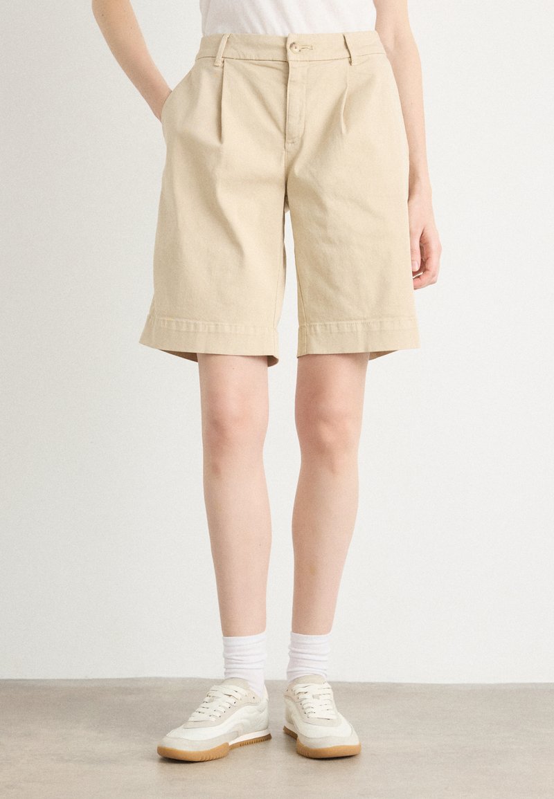 Shorts en coton beige avec des plis, des ourlets retroussés, des poches latérales et une fermeture à bouton à l'avant, portés avec des baskets blanches et des chaussettes de cheville.