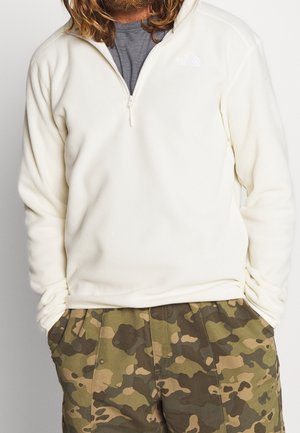 Mann trägt cremefarbenen Fleece mit halbem Reißverschluss und grün-braune Camouflagehose mit Händen in den Taschen, beschnitten unterhalb der Schultern.