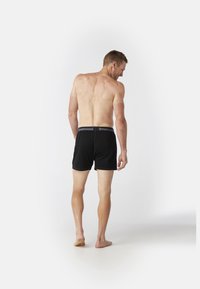 Zwarte boxershorts van wol, met een gebrandmerkt elastiek. Het model is van achteren bekeken, met een neutrale achtergrond en blote voeten.