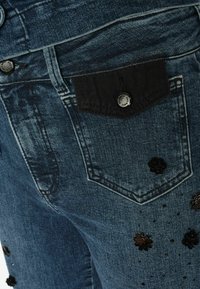 Modré džínsy vyrobené z denimu s tmavým látkovým vreckom, dekoratívnymi čiernymi kvetinovými aplikáciami a jemným zrelaxovaním v oblasti pásu.