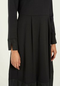Schwarzes Kleid mit langen Ärmeln, das mit strukturiertem Stoff und satinähnlichen Akzenten an den Bündchen, einem plissierten Design und einer fließenden Silhouette versehen ist.
