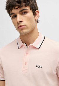 Maglietta polo rosa chiaro con colletto a righe nere e bianche, tessuto testurizzato, chiusura con tre bottoni e logo "BOSS" nero sul petto.