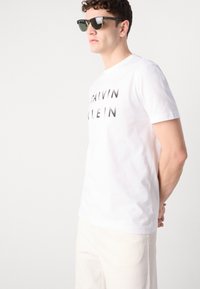 White cotton T-shirt featuring a bold black "CALVIN KLEIN" print. Paired with dark sunglasses, model stands with hands behind back.