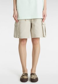 Beige cargoshorts med fransat fåll, med två sidofickor. Bärs med bruna mönstrade slip-on skor. Enkelt, avslappnat design.