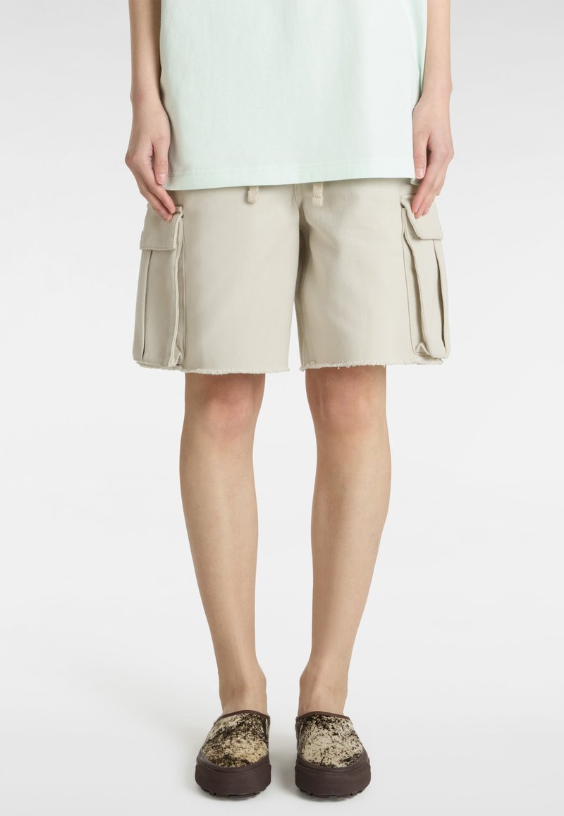 Beige cargoshorts med fransat fåll, med två sidofickor. Bärs med bruna mönstrade slip-on skor. Enkelt, avslappnat design.