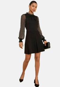 Black dress with a fitted bodice and pleated skirt, featuring polka dot sheer sleeves and a ruffled collar. Worn with black heels and a clutch.