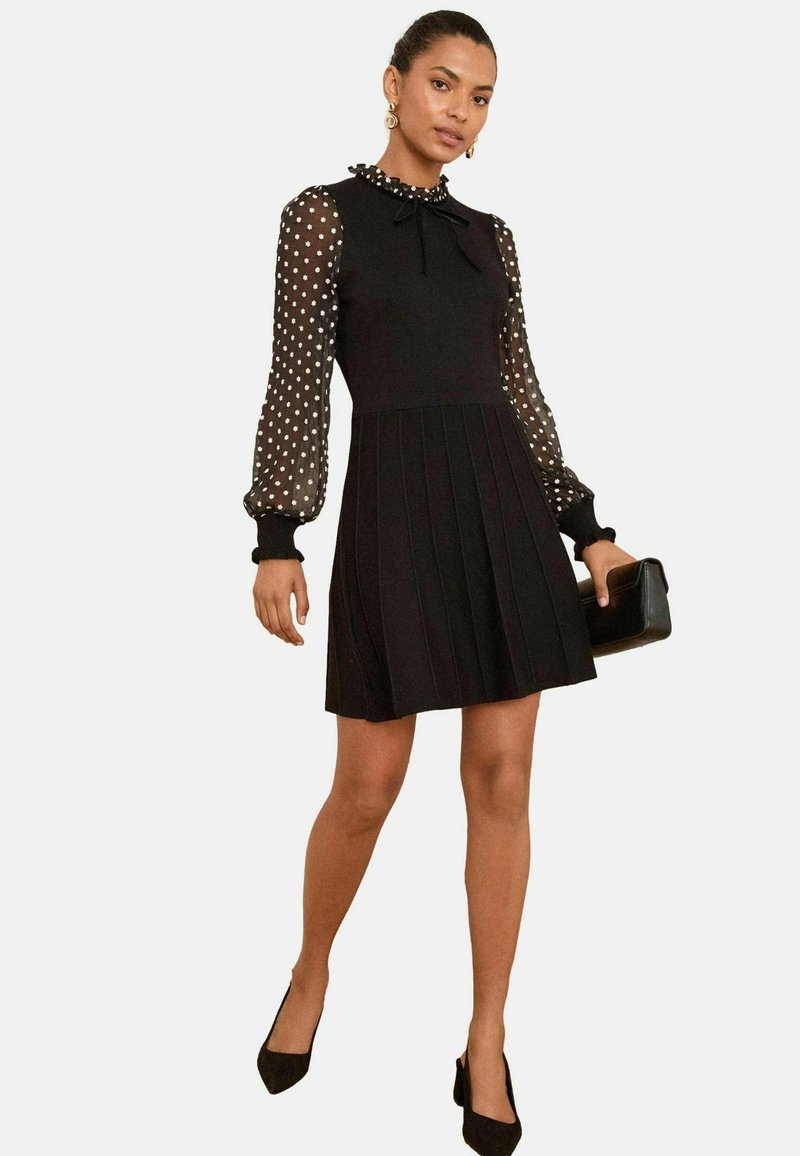 Black dress with a fitted bodice and pleated skirt, featuring polka dot sheer sleeves and a ruffled collar. Worn with black heels and a clutch.