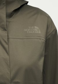 Olívazöld kapucnis dzseki, amelyen a „The North Face” logó dombornyomottan jelenik meg a felső mellrészen, és a dzseki elején varrásrészletek láthatók.