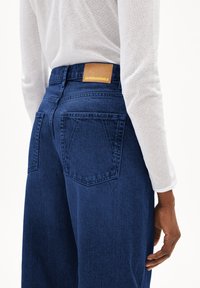 Donkerblauwe spijkerbroek met wijde pijpen, voorzien van twee achterzakken en een tan leren merkpatch op de taille. De stof heeft een gladde textuur.