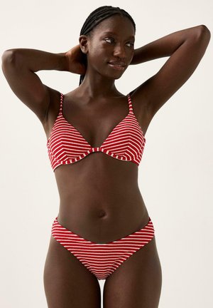 Kvinde med flettet hår iført en rød og hvidstribet bikini, poserer med hænderne bag hovedet mod en ensfarvet baggrund.