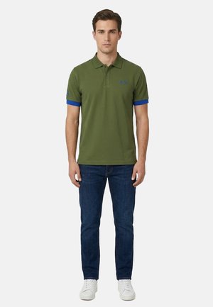 Uomo in piedi che indossa una polo verde oliva con bordi blu, jeans blu scuro e scarpe da ginnastica bianche su uno sfondo chiaro uniforme.