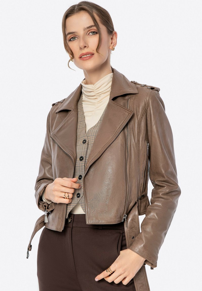 Braune Lederbikerjacke mit glänzender Oberfläche, Reverskragen, Reißverschlussdetails und asymmetrischem Schnitt. Über einem beigen Rollkragenpullover getragen.