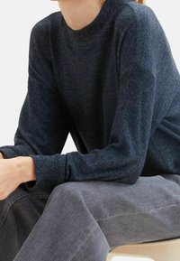 Tummansininen neulepaita, jossa on kuvioitu, neulottu kangas ja resorikaulus. Mallin kädet lepäävät vaaleanharmaiden housujen päällä.