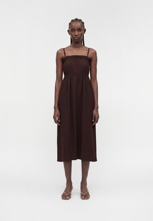 Femme aux cheveux tressés portant une robe midi marron foncé sans manches et des talons beige à brides, debout devant un fond clair uni.