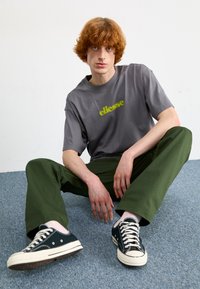 Grijze t-shirt met korte mouwen en logo "ellesse" in het geel, gecombineerd met groene casual broek en zwarte sneakers. Achtergrond is een lichte kleur.