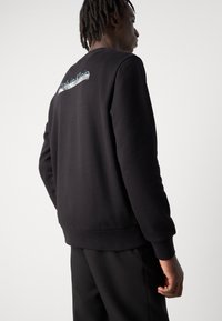 Zwarte sweatshirt met ribbelmanchetten en -zoom, voorzien van een wit en iriserend "Calvin Klein" logo op de bovenrug. Glad textuur.