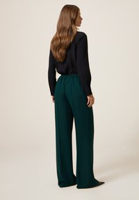 Blusa negra de manga larga combinada con pantalones anchos verdes. Los pantalones cuentan con una cintura elástica; la modelo tiene el cabello largo y ondulado.