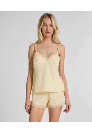 Femme portant un caraco jaune pâle orné de dentelle et un short assorti, debout devant un fond uni gris clair.