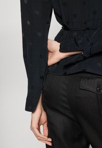 Černá blůza s puntíky, s hladkým povrchem a mírným leskem. Vyznačuje se přiléhavým pasem a úzkými rukávy. Vzorem se hodí k texturovaným černým kalhotám.