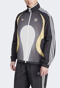 Vīrietis valkā melnu un bēšu Adidas sporta jaku ar baltām trim svītrām uz piedurknēm un atbilstošas melnas bikses ar NTS logotipu.