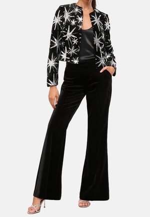 Vrouw draagt een zwart fluwelen jasje met zilveren sterrenpatronen, een zwarte satijnen top, zwarte fluwelen broek met wijde pijpen en zilveren sandalen met hoge hakken.