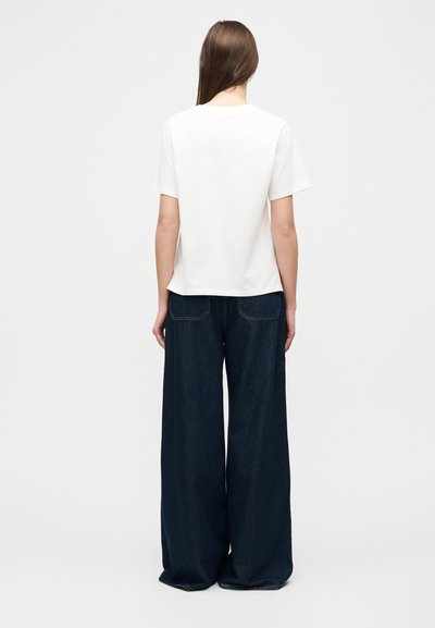 Γυναίκα που στέκεται γυρισμένη με την πλάτη, φορώντας ένα απλό λευκό t-shirt και σκούρο μπλε τζιν με φαρδιά πόδια, μπροστά σε απλό φωτεινό φόντο.
