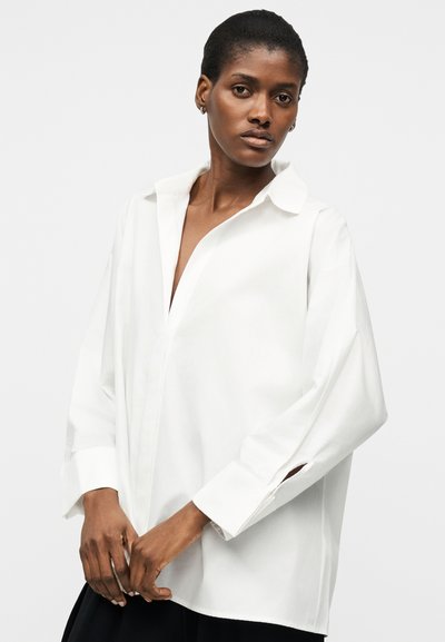 Žena s krátkými vlasy, oblečená v volné bílé košili na knoflíky a černých kalhotách, stojící před jednoduchým světlým pozadím.