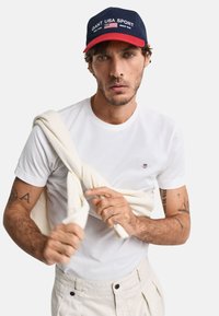 Hvid kortærmet t-shirt, lys beige bukser og en marineblå hat med rød skygge, der har teksten "GANT USA SPORT" og et amerikansk flag emblem.