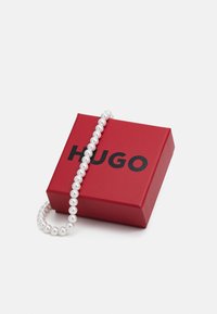 Piros ajándékdoboz, rajta fekete "HUGO" felirattal, benne egy fehér gyöngyökből készült nyaklánc, sima, fényes felülettel bemutatva.