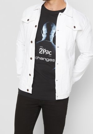 Mann trägt ein schwarzes 2Pac "Changes" Grafik-T-Shirt unter einer offenen weißen Jacke mit Knöpfen und schwarzen Hosen, steht mit hinter dem Rücken verschränkten Händen.
