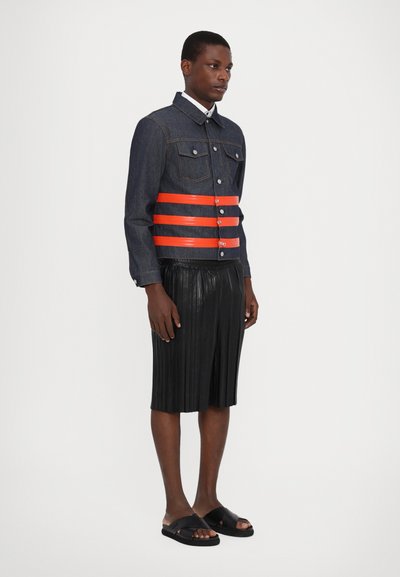 Veste en jean avec des détails rayés orange, short plissé noir et sandales noires. Les caractéristiques clés incluent des fermetures à boutons et des poches.
