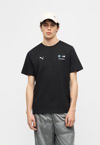 Νέος άνδρας που φοράει μαύρο t-shirt BMW Motorsport Puma, γκρι παντελόνι και λευκό καπέλο Puma, στέκεται μπροστά σε απλό φόντο.