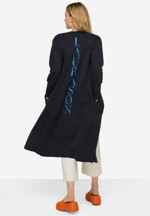 Lange marineblauwe jas met een verticale turquoise grafische tekst op de rug, voorzien van zijzakken en een relaxed fit, in combinatie met oranje plateauschoenen.