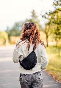 Černá měkká taška přes rameno s křivkovým tvarem, texturovaným povrchem a zipem, nošená přes světle šedou mikinu a roztrhané džíny.