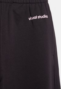 Pantaloni sport negri cu talie elastică. Prezintă un logo roz subtil "visual studies" brodat în față. Textură a țesăturii netedă.