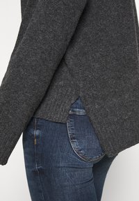 Suéter gris de punto con textura acanalada y aberturas laterales, combinado con unos pantalones vaqueros de mezclilla azul oscuro que presentan un diseño clásico de bolsillo.