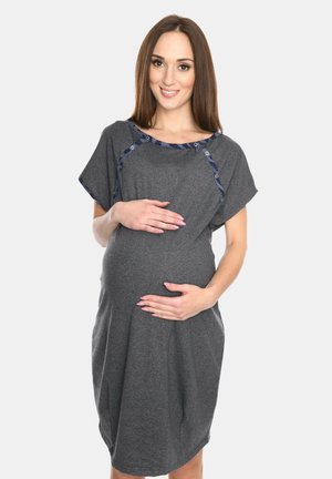 Schwangere Frau in einem grauen Umstandskleid hält sanft ihren Bauch und lächelt in die Kamera vor einem einfarbigen Hintergrund.